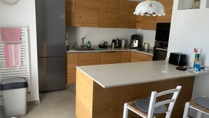 Een moderne keuken met houten kastjes, een roestvrijstalen koelkast, een koffiezetapparaat en een broodrooster. Er staan twee witte stoelen met gevlochten zittingen.