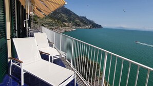 Ein Balkon mit weißen Stühlen und einem gestreiften Sonnensegel, das auf eine Küstenlandschaft mit klarem, blauem Meer und fernen Bergen blickt.