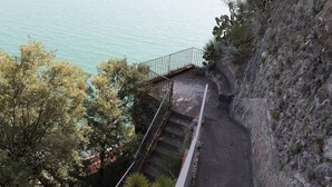 Eine Treppe führt hinab zu einem Küstenpfad mit Geländer, umgeben von Bäumen und felsigem Gelände.