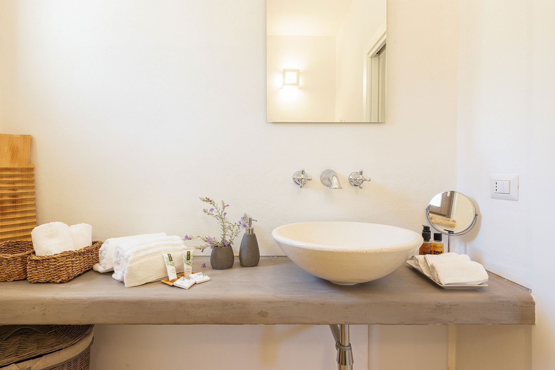 A bathroom with a large white sink, a mirror, and a wall-mounted light. There are towels and toiletries on the counter.