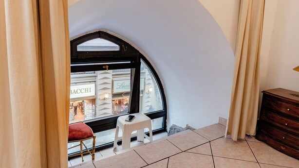 A room with a curved wall, a window with a view of a store, a white stool, and a wooden dresser. - Amedei Apartment: 2 suites {10 m from Ponte Vecchio} (Firenze)