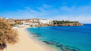 Une plage côtière aux eaux bleues et limpides, avec un rivage sablonneux et des bâtiments blancs en arrière-plan.