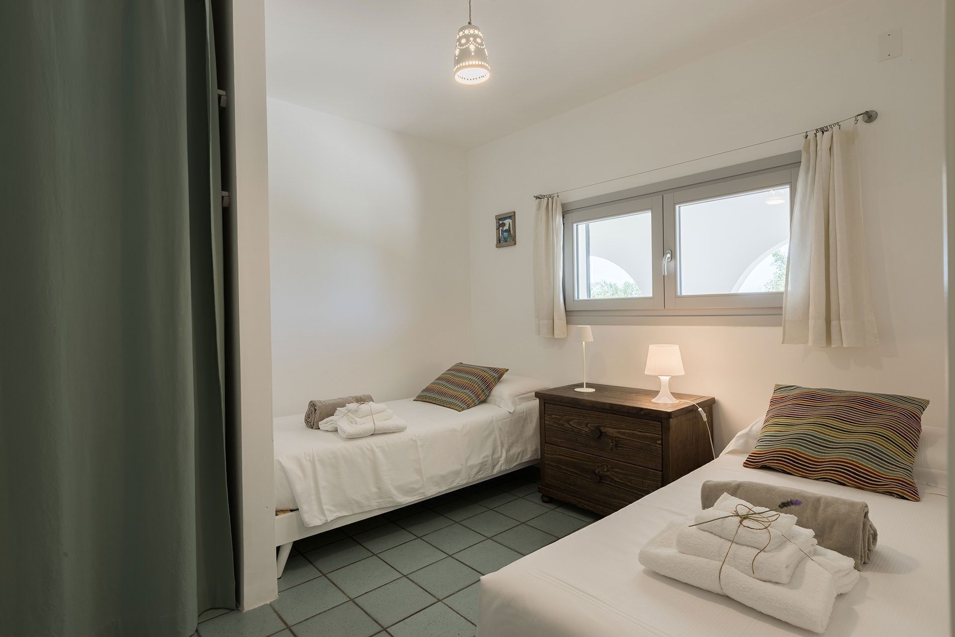 A small, clean room with two beds, a window, and a bedside table with a lamp.
