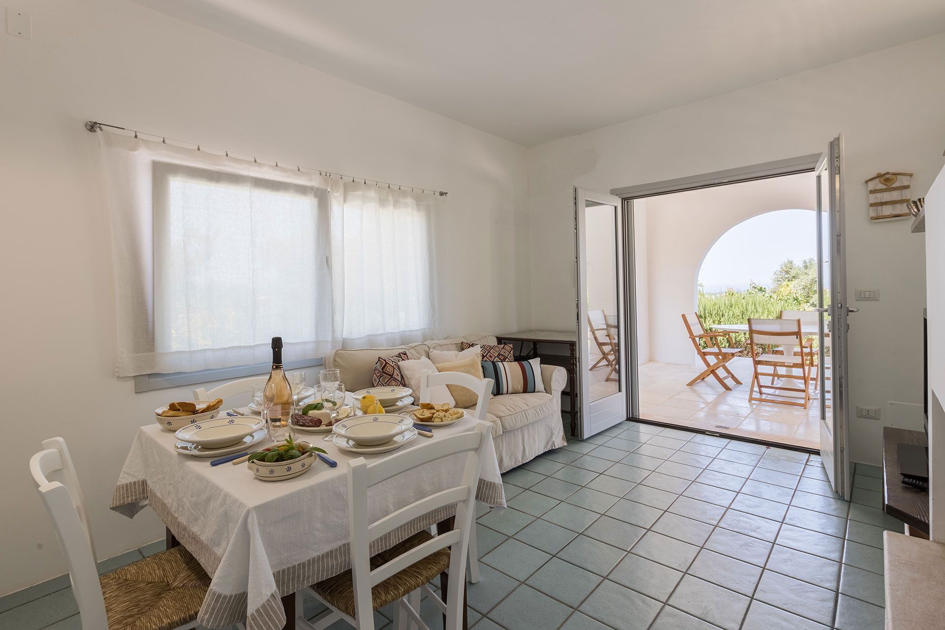 Ein Essbereich mit Tisch für vier Personen, einer Couch und Blick durch eine offene Tür auf eine Terrasse.