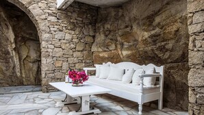 Un sofá blanco con cojines, una mesa blanca y un jarrón con flores rosadas en un espacio al aire libre con paredes de piedra.