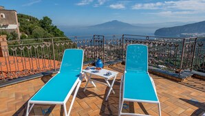 Dos sillones azules con una mesa y un jarrón en una terraza con vistas a una ciudad y montañas.