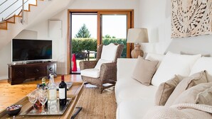 Una sala de estar moderna con televisor, una silla de mimbre, un sofá blanco y una zona de comedor con una bandeja de botellas y vasos.