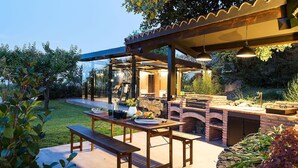 Un patio exterior con mesa y banco de madera, una barbacoa de ladrillo y una pérgola con luces colgantes.