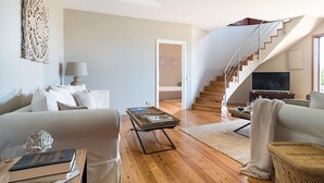 Amplia sala de estar con sofá, una mesa de centro y una estantería. Una escalera conduce a un nivel superior con un televisor.