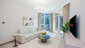 A modern living room with a white sofa, a coffee table with flowers, a large window with curtains, and a flat-screen TV mounted on the wall. - Stylish 2BR Fully Furnished Unit at Business Bay (Dubai)
