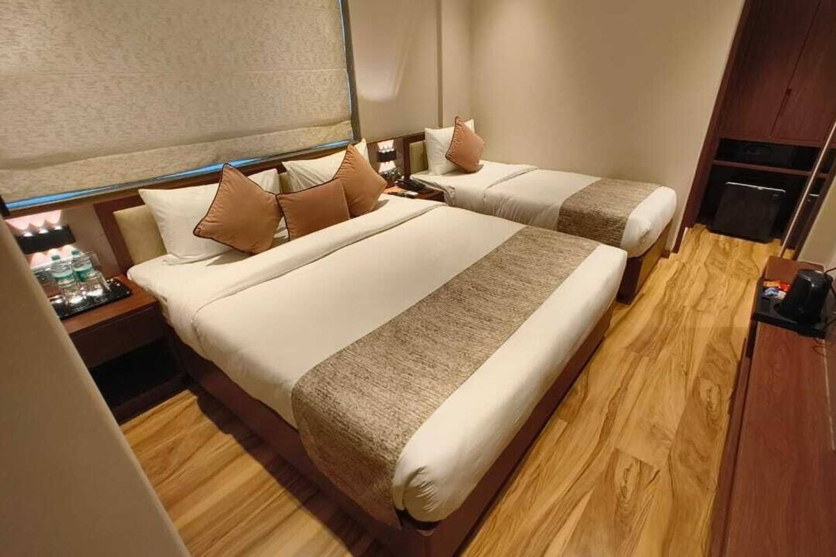 A hotel room with two beds, wooden flooring, a nightstand with a lamp, and a small table with a coffee maker.