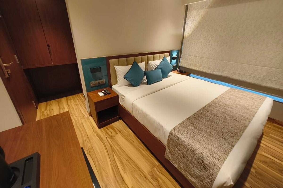 A modern hotel room with a wooden floor, a bed with white and teal pillows, a nightstand, and a large window with a view.