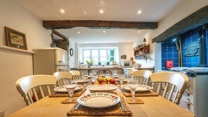A dining area with a wooden table set for four, surrounded by chairs, with a kitchen in the background. - 4Bed Thatched Cottage in Oxfordshire (Ledwell)