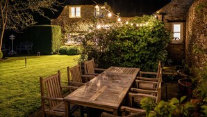 An outdoor wooden table and chairs set in a garden at night, surrounded by stone walls and string lights. - 4Bed Thatched Cottage in Oxfordshire (Ledwell)
