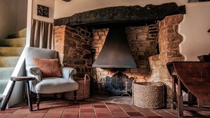 A cozy room with a stone fireplace, a comfortable armchair, and a wooden staircase. - 4Bed Thatched Cottage in Oxfordshire (Ledwell)