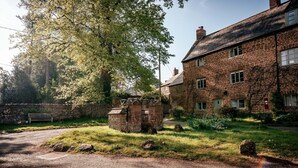 A traditional stone building with a thatched roof, surrounded by a stone wall and a grassy area with a small pond. - 4Bed Thatched Cottage in Oxfordshire (Ledwell)