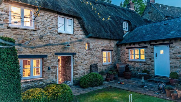 A stone house with multiple windows, a thatched roof, and a small garden with potted plants and a lawn. - 4Bed Thatched Cottage in Oxfordshire (Ledwell)