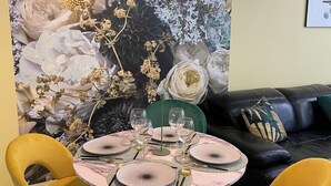 Un comedor con una mesa redonda dispuesta para cuatro personas, rodeado por sillas amarillas, un sillón tapizado en verde y un sofá de cuero negro con cojines estampados.