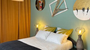 Un dormitorio moderno con cama, dos lámparas de noche, una obra de arte geométrica en la pared y un amplio ventanal con cortinas.