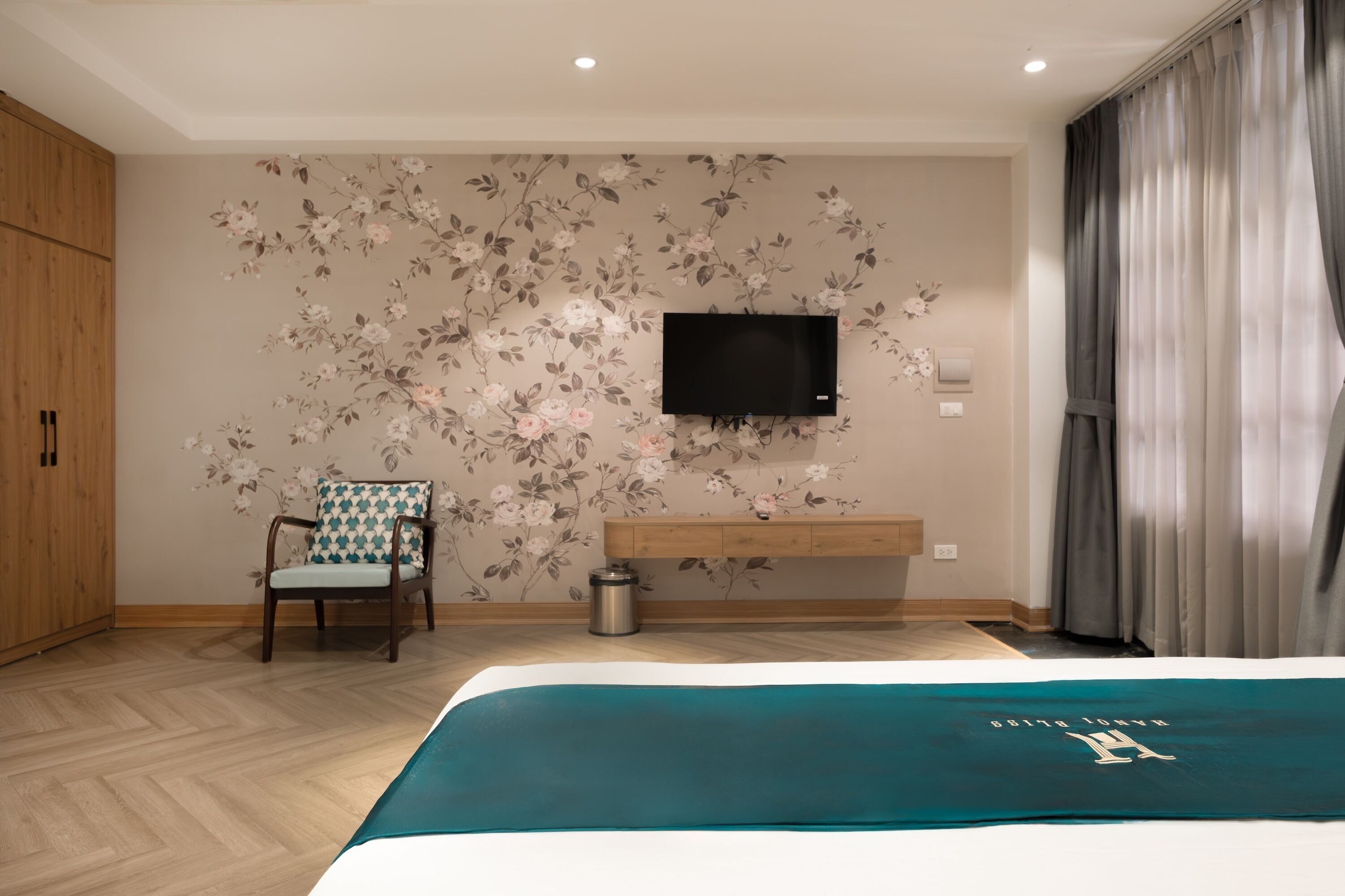 A hotel room with a bed, a TV mounted on the wall, a chair, a wooden cabinet, and a trash bin.