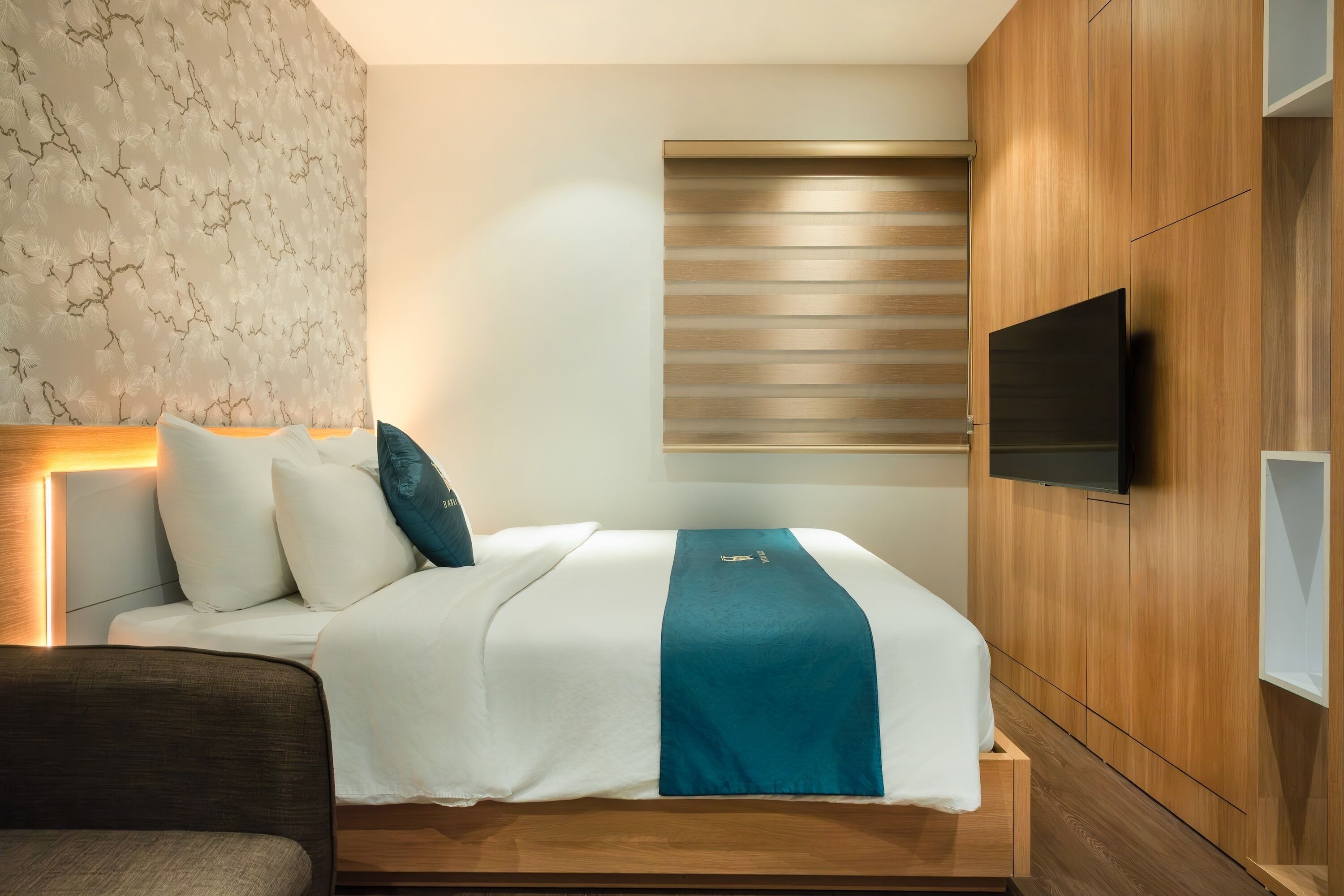 A modern hotel room with a neatly made bed, a flat-screen TV mounted on the wall, and a wooden wardrobe.