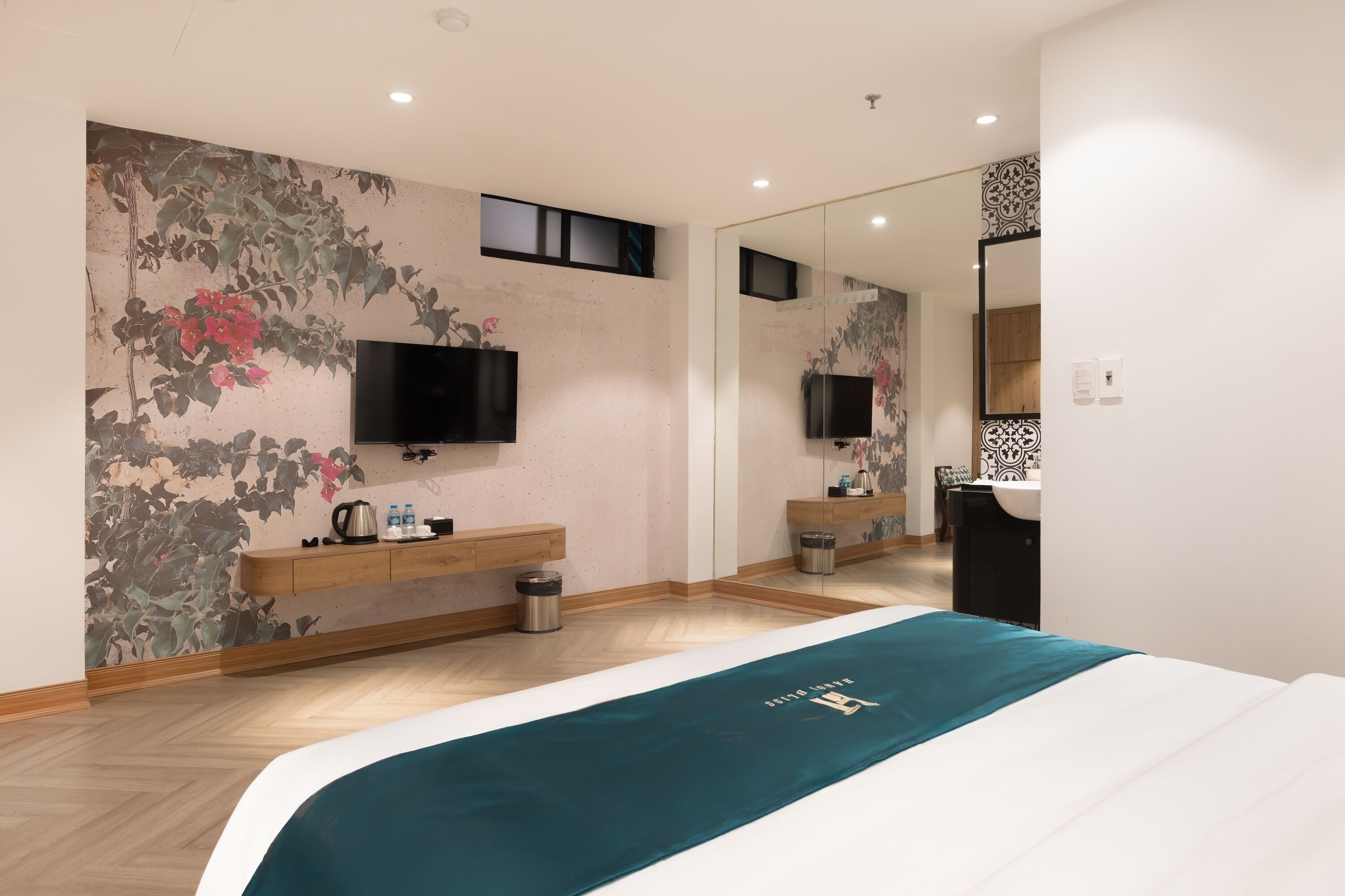 A modern hotel room with a large bed, a flat-screen TV mounted on the wall, a wooden nightstand, a mirror, and a bathroom with a sink and toilet.