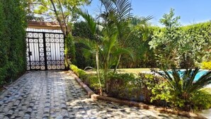 A paved pathway leading to a gated entrance surrounded by lush greenery and a swimming pool.