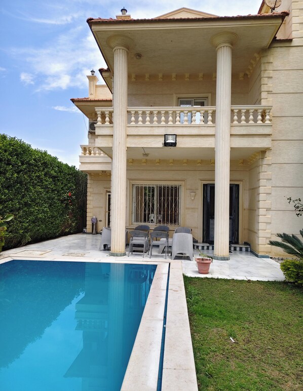 Una casa de dos pisos con piscina, mobiliario exterior y un jardín bien cuidado.