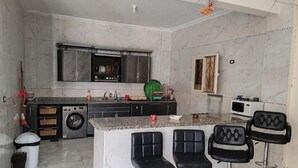 A kitchen with a marble countertop, a washing machine, and a microwave. There are three black bar stools in front of the counter.