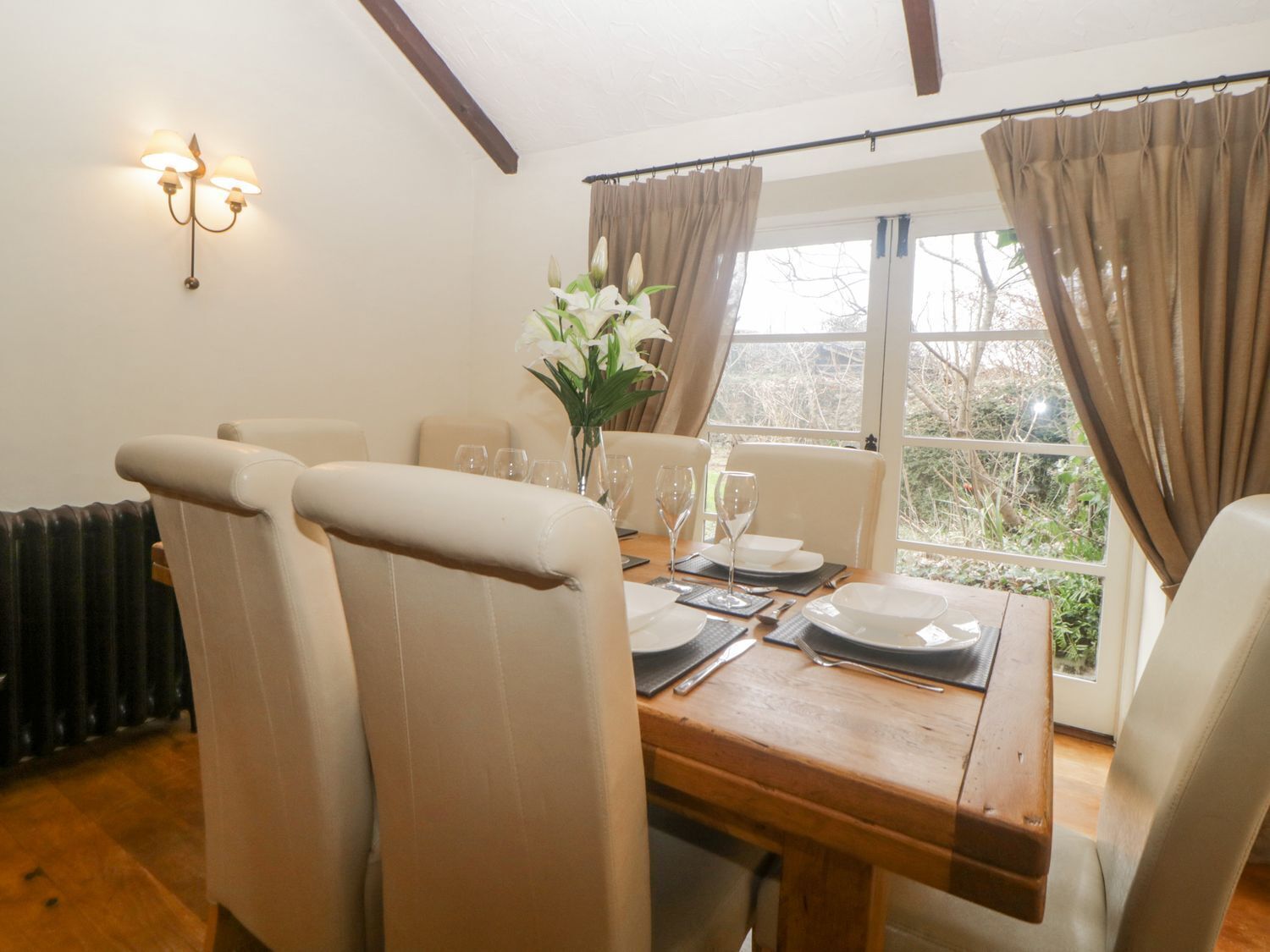 Een eetkamer met een houten tafel gedekt voor vier personen, beigekleurde stoelen, een vaas met witte bloemen en een raam met gordijnen.