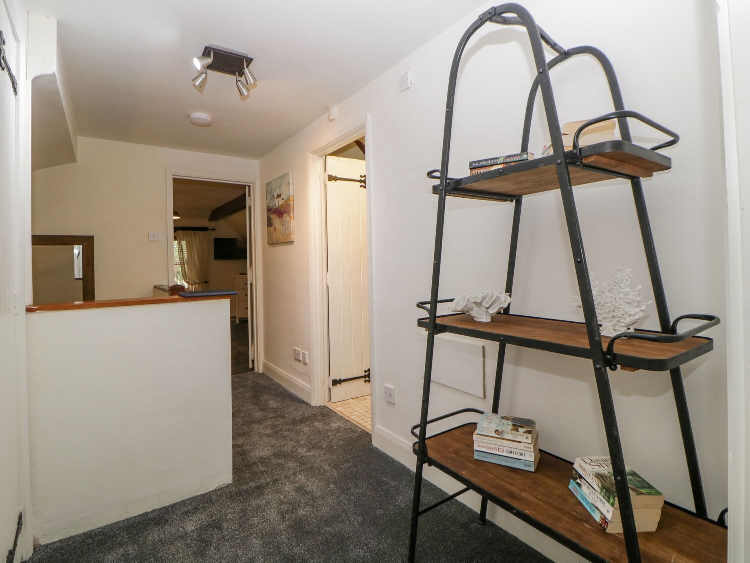 A multi-tiered metal shelf unit with books and decorative items, positioned in a hallway with carpeted flooring and white walls.