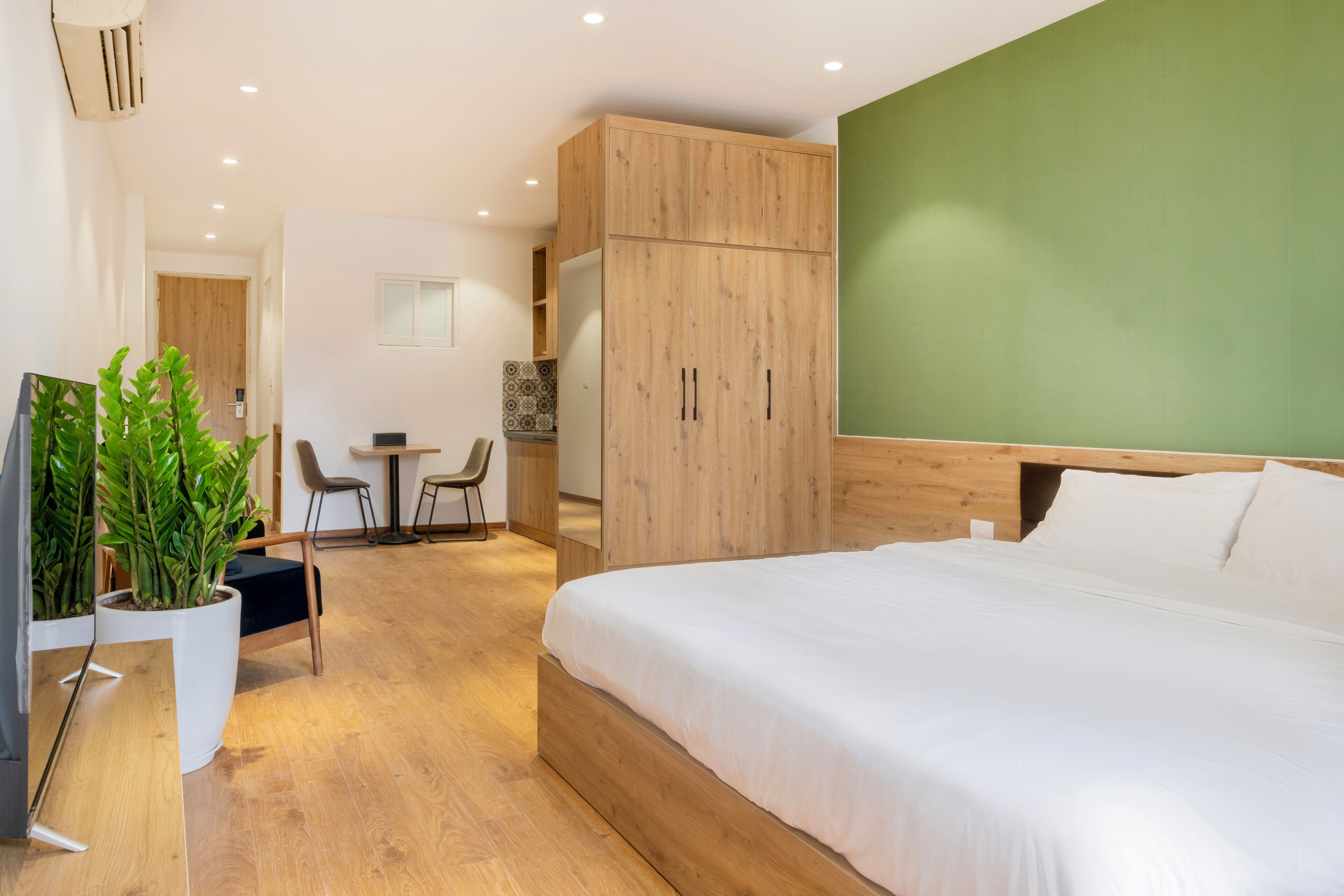 A modern hotel room with a wooden bed, a green accent wall, a dining area, and a TV.