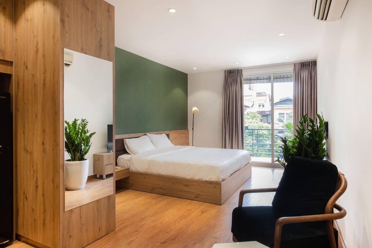 A modern bedroom with a wooden bed, a nightstand, a potted plant, a chair, and a large window with curtains.