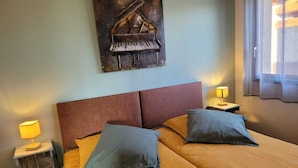 A bedroom with a bed, two pillows, a headboard, a nightstand with a lamp, and a painting of a piano on the wall.