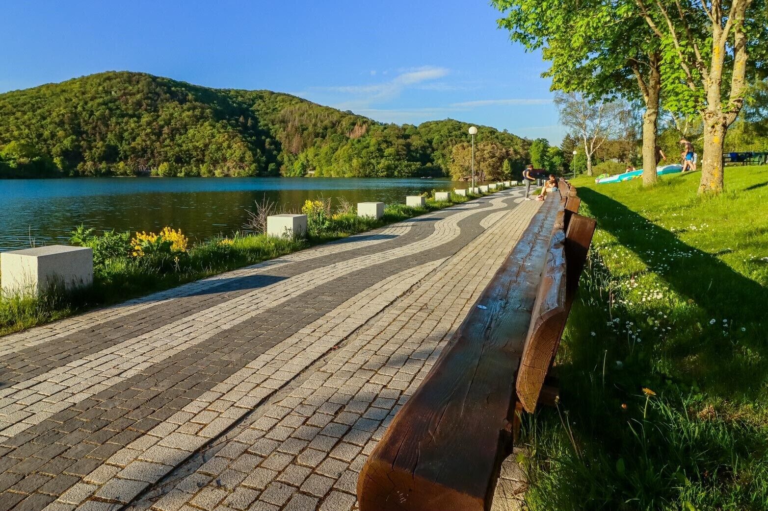 A paved pathway alongside a body of water with trees and a hilly landscape in the background.