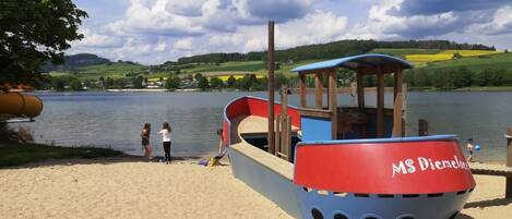 A boat with "MS Dierkelse" written on it, docked at a sandy beach with a lake and hills in the background.