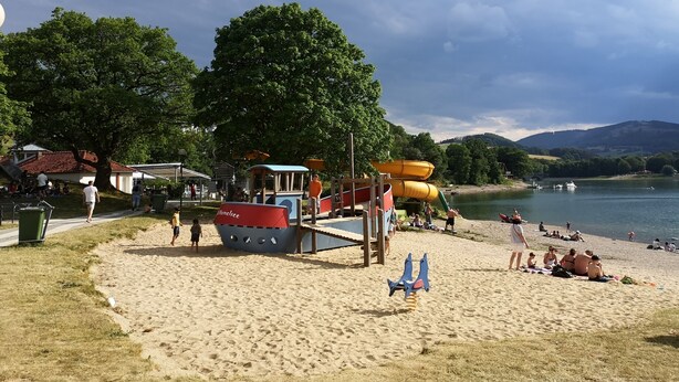 A beach with a playground, including slides and swings, and a lake with boats in the background.