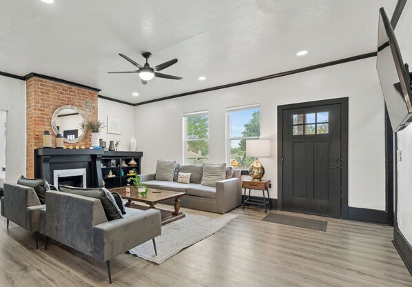 A spacious living room with a fireplace, a ceiling fan, and a large window.