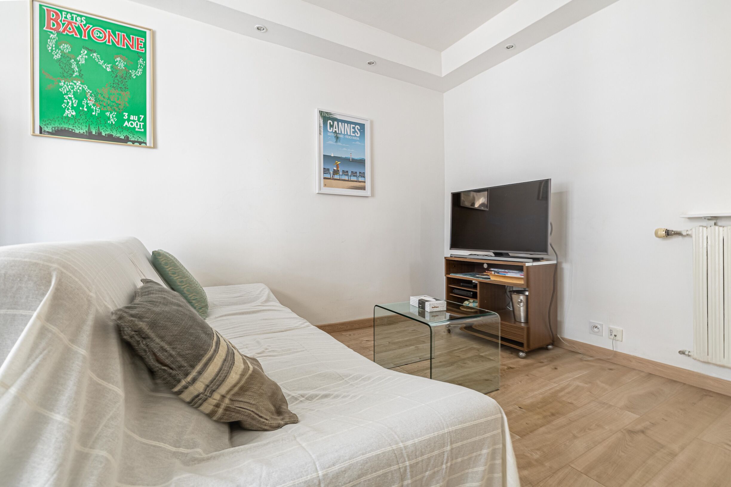 A living room with a sofa, a glass coffee table, a TV on a stand, and two posters on the wall.