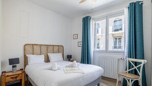 En pænt redt seng med hvide sengetøj, et natbord med lampe, en stol og et vindue med gardiner.