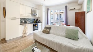 En kompakt opholdsstue med et tekøkken, en sofa med hynder, et glasbord og en ventilator i loftet.
