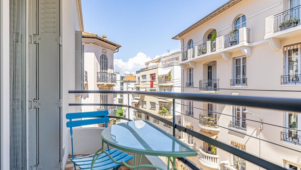 En balkon med et glasbord og blå stole, der giver udsigt over en række hvide bygninger med balkoner.