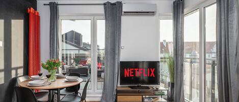A modern living room with a dining area, large windows, and a TV displaying the Netflix logo.