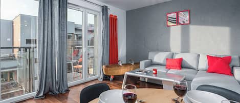 A modern living room with a dining table set for two, a grey sofa with red cushions, and a large window with grey curtains.