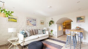 Una sala de estar moderna con un sofá blanco, un otoman redondo y un tapete a rayas. Se observan cuadros enmarcados en las paredes, una planta colgante y una mesita de noche con una lámpara.