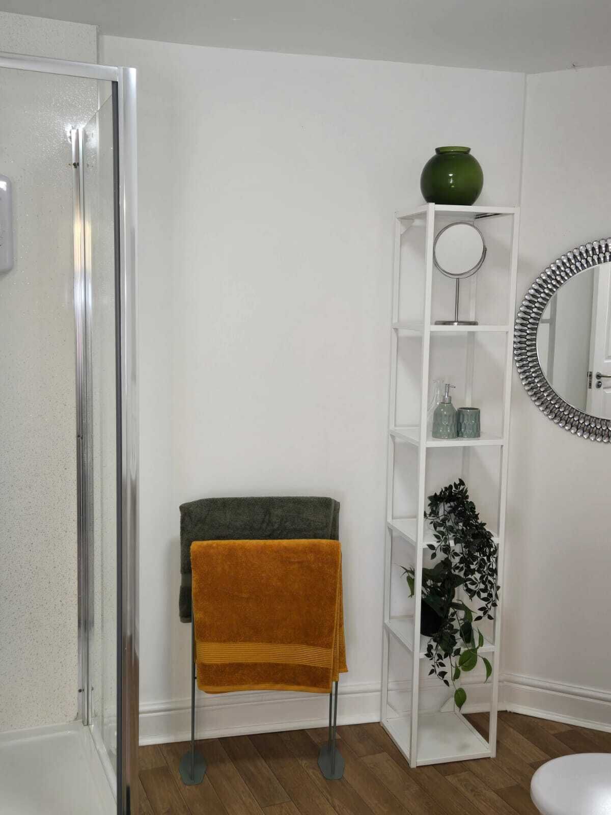 A bathroom with a glass shower enclosure, a white vanity with a green vase, a mirror with a decorative frame, and a shelf with toiletries.