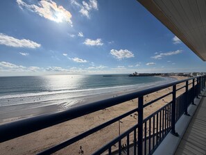 A beach view with a clear sky, a sandy shore, and a promenade with a railing.