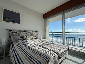 A bedroom with a large bed, a headboard with a nautical-themed print, a bedside table, and a window with a view of the sea.