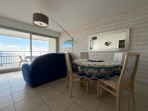 A room with a round table, chairs, a sofa, a large window, and a view of the sea.