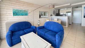 Una sala moderna con dos sofás azules, una mesa de centro blanca, una lámpara de pie, un cuadro y una cocina al fondo.